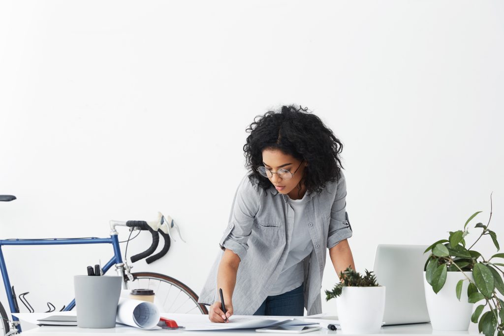 portrait of successful woman engineer having dark curly hair wearing casual shirt and eyeglasses leaning at white table signing important business documents isolated over cosy office interior